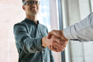 A smiling man in glasses shakes hands with another person in a bright office setting, symbolizing a successful agreement or consultation.