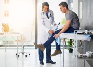 A middle-aged male patient sits on an exam table while a doctor in a white coat examines his injured leg inside a modern medical office.
