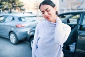 A woman standing near a car accident scene, holding her neck in pain, indicating a possible whiplash injury following a rear-end collision.
