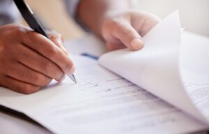 A person signing a legal document with a pen, close-up view of hands and paperwork, symbolizing agreement or legal proceedings.