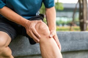 A person in athletic shorts sits on an outdoor bench, gripping their knee in visible discomfort, indicating knee pain or injury.