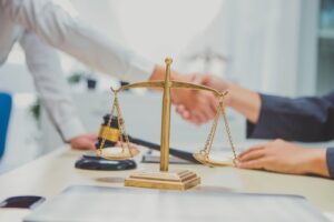 Two individuals shake hands behind a golden balance scale and gavel on a desk, representing a legal agreement or settlement in a law office.