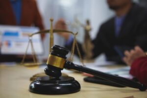 Close-up of a judge’s gavel and scales of justice on a courtroom table, with blurred figures in business attire discussing legal matters in the background.