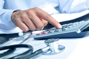 Close-up of a doctor's hand pressing buttons on a calculator with a stethoscope and medical paperwork on the desk, symbolizing medical billing or healthcare cost calculation.