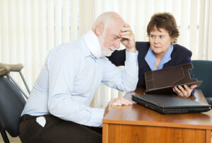 Senior man wearing a neck brace looks distressed while speaking with a concerned female attorney, both seated at a desk with legal documents and a laptop.