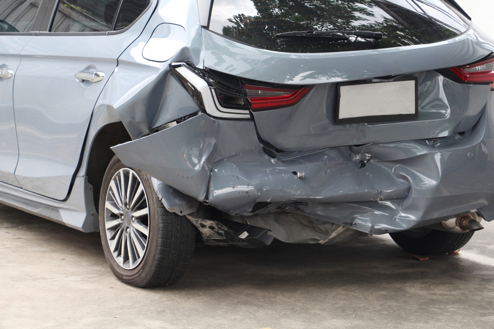 Rear view of a grey car with significant damage to the back bumper and trunk area after a rear-end collision, parked in a garage.
