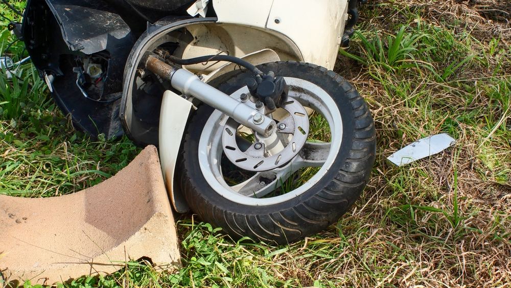 Close-up of a damaged white scooter lying on its side in grass after a crash, with the front wheel twisted and debris scattered nearby.