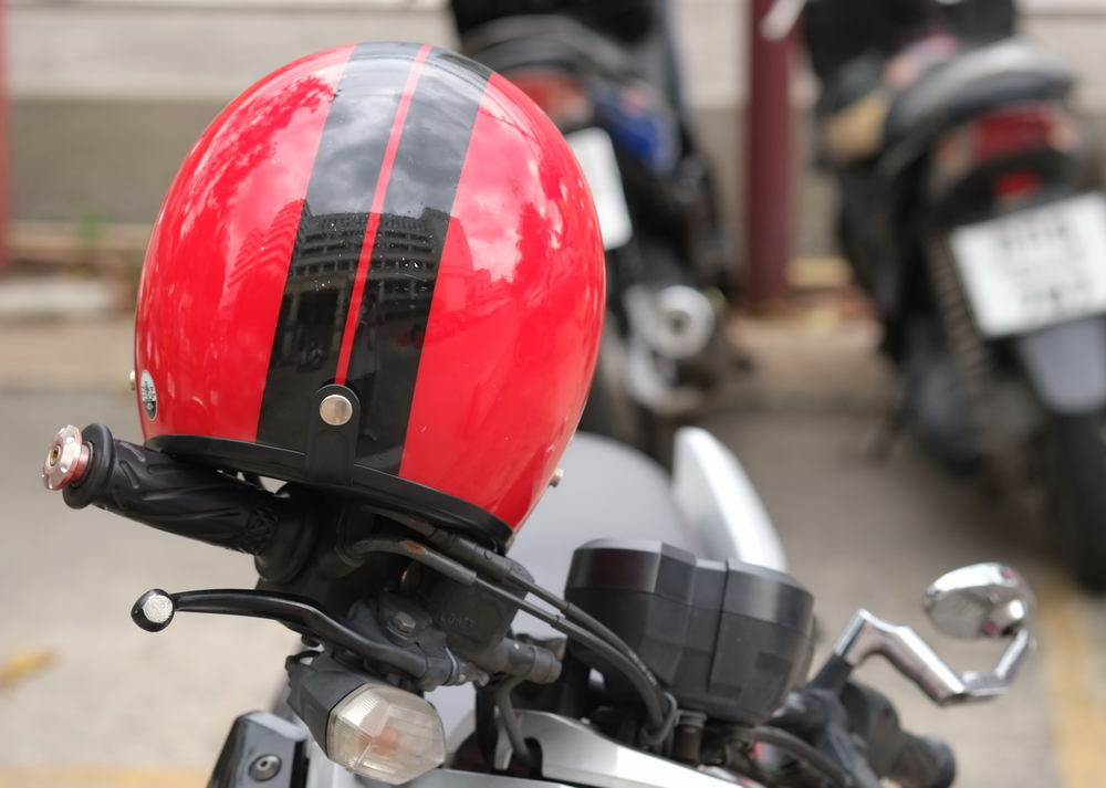 Close-up of a red motorcycle helmet with a black racing stripe resting on the handlebars of a parked motorcycle, with other motorcycles blurred in the background.