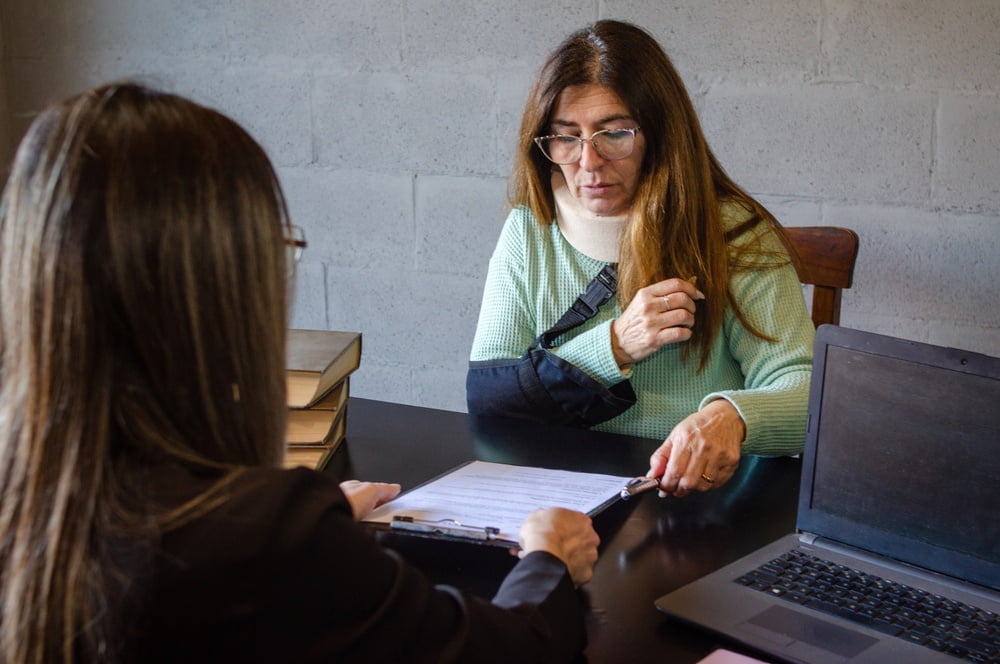 Person with an arm sling and neck brace meeting with an attorney to review legal documents.